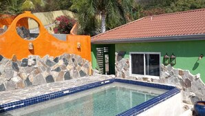 Une piscine avec des murs en pierre et un bord carrelé, un bâtiment écologique avec un toit rouge, et un mur en pierre percé d’une ouverture circulaire.