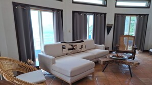Un salon avec un canapé blanc, un fauteuil en osier, une table basse en bois et une vue sur l’extérieur grâce à de grandes fenêtres ornées de rideaux.
