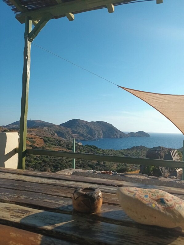 A terrace with a view of the sea and mountains, a wooden table with a ceramic pot and a decorative item, and a shade sail.