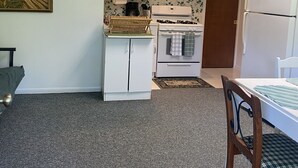 A compact kitchen with white cabinets, a stove, and a refrigerator. There is a dining table with a chair, a small shelf with glassware, and a painting on the wall. - CURRENTLY UNDER REPAIR (Deersville)