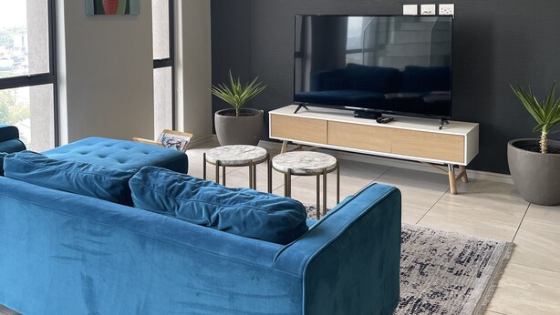 A modern living room with a blue sofa, a flat-screen TV, two small round tables, and potted plants. - CGRSA Apartment Sandton (Sandton)