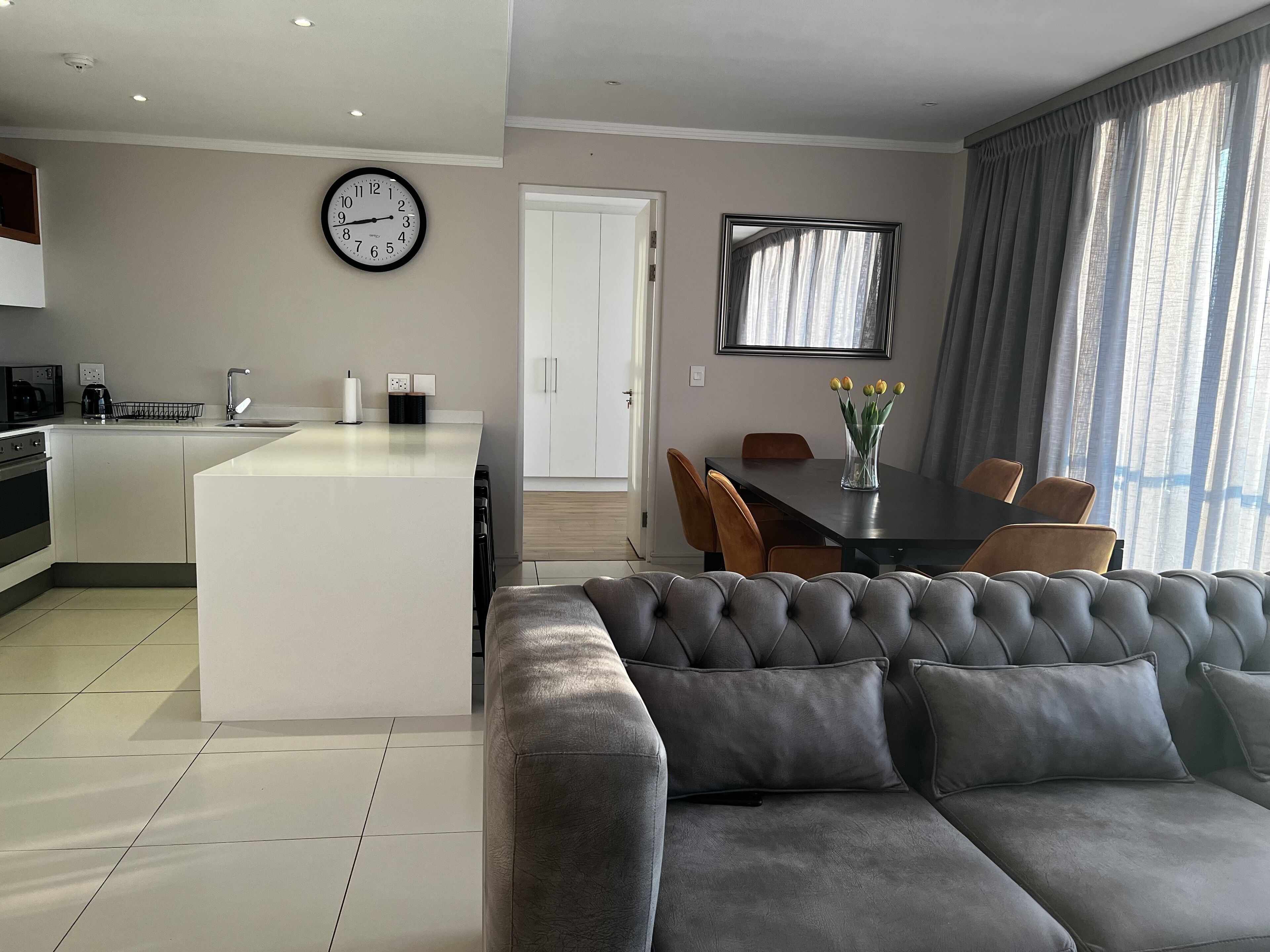 Un espacio habitable moderno con un sofá gris, una zona de comedor con mesa y sillas, una cocina con una isla blanca, un reloj en la pared y un amplio ventanal con cortinas.