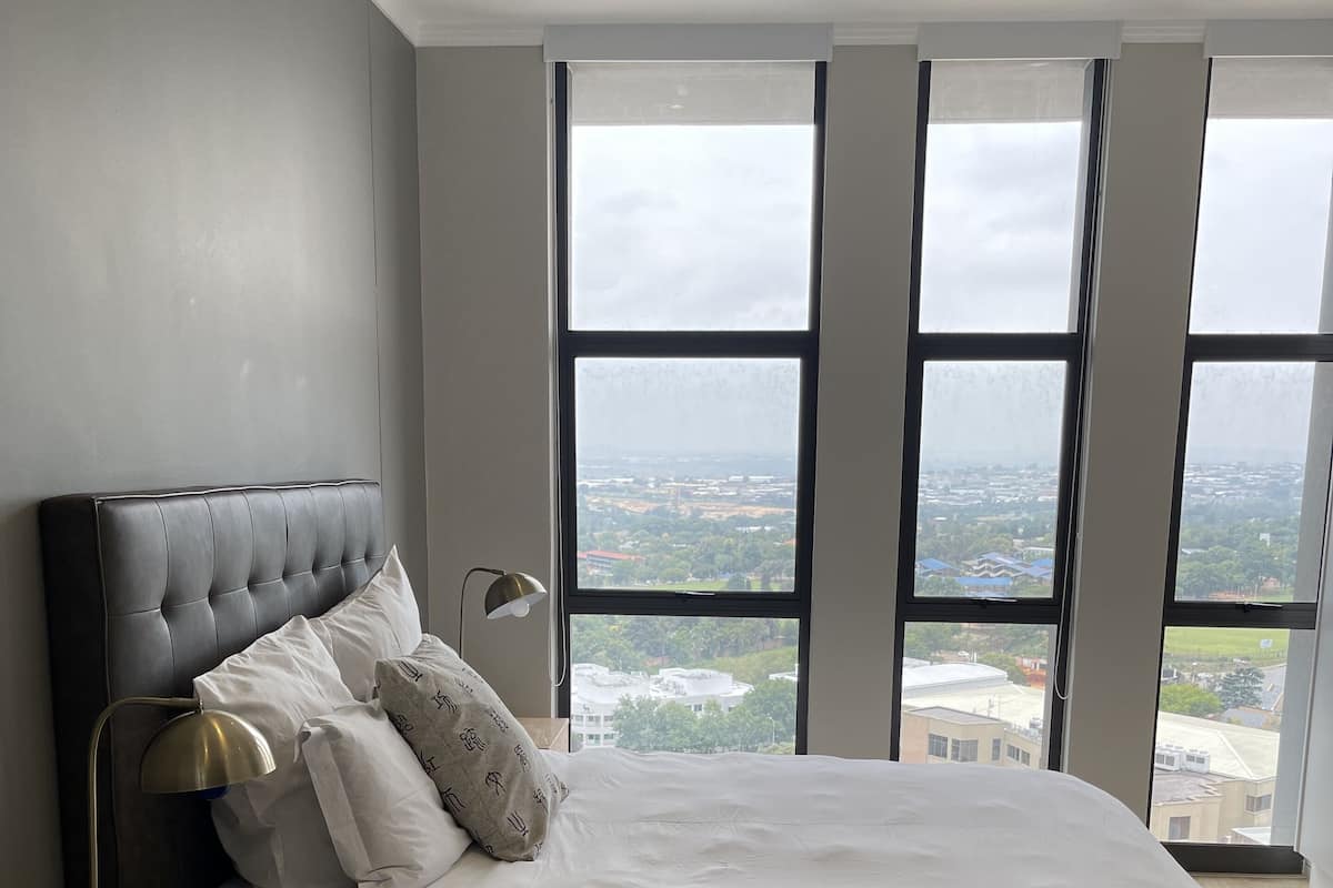 A bedroom with a large bed, a nightstand, a lamp, and a view of a cityscape through large windows.