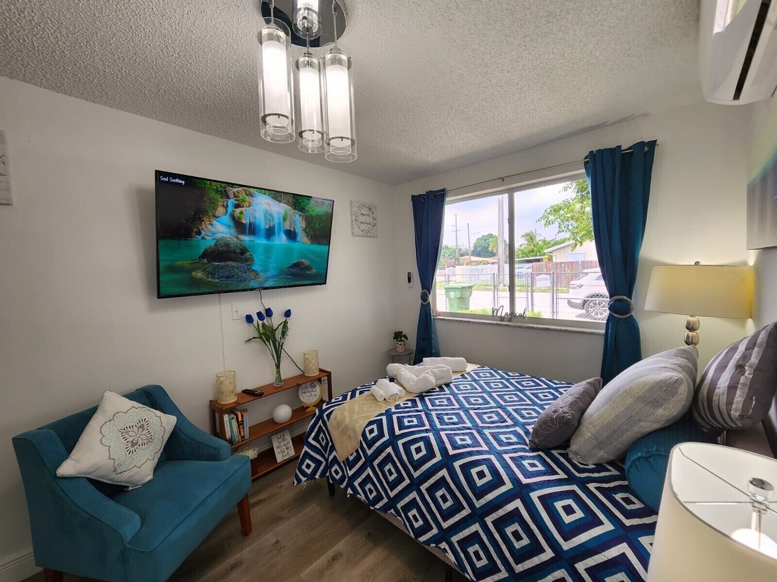 A bedroom with a bed, a blue armchair, a TV on the wall, and a window with curtains.