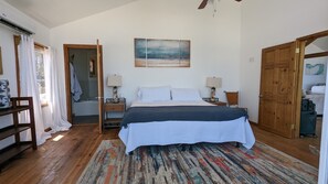 A bedroom with a bed, two bedside tables, a window with curtains, a wooden door, and a shower area.