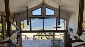 A spacious room with large windows overlooking a beach, wooden beams, a ceiling fan, and a wooden deck. - Spacious Beach Front Home  -  Swim, Snorkel, Dive, Paddle, at your doorstep! (Sandy Bay)