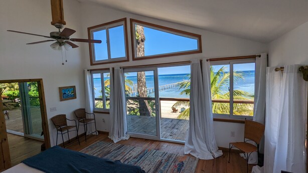 A room with large windows overlooking a beach, a ceiling fan, and a bed with a blue blanket. - Spacious Beach Front Home  -  Swim, Snorkel, Dive, Paddle, at your doorstep! (Sandy Bay)