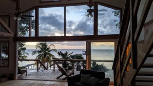 A spacious room with large windows overlooking a beach, a wooden deck, and a view of the ocean. - Spacious Beach Front Home  -  Swim, Snorkel, Dive, Paddle, at your doorstep! (Sandy Bay)