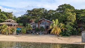 A beachfront property with a large wooden house, a thatched-roof structure, and a sandy beach. - Spacious Beach Front Home  -  Swim, Snorkel, Dive, Paddle, at your doorstep! (Sandy Bay)