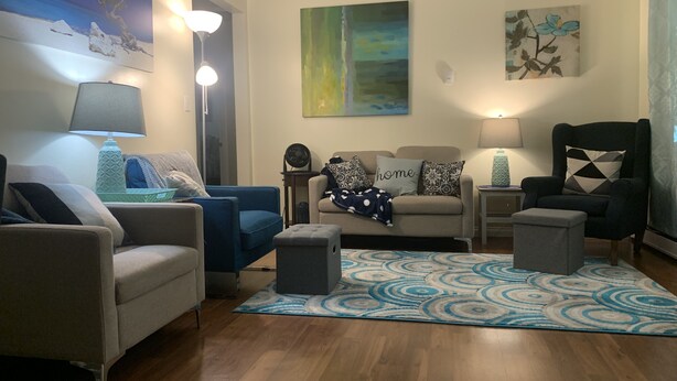 A living room with a sofa, armchairs, a coffee table, and a rug. There are paintings on the walls and a lamp on the side table. - Relaxing - Two Bedroom Suite in Red Deer (Red Deer)