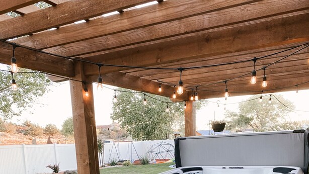 Un bain à remous sous une pergola en bois, agrémentée de guirlandes lumineuses, une table avec des chaises et une cour arrière verdoyante.