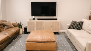 Un salon doté d’un grand téléviseur à écran plat fixé au mur, d’un canapé en cuir marron, d’un canapé beige et d’un petit pouf placé au centre.