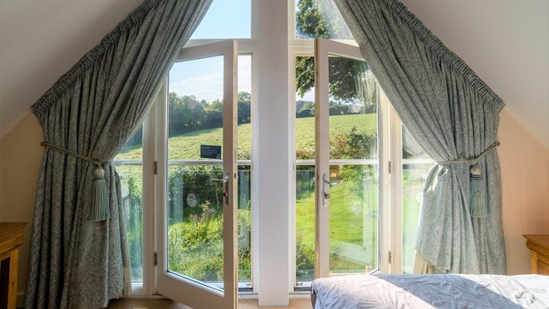 Ein Schlafzimmer mit einem großen Dachfenster und Vorhängen, einem Bett sowie Blick auf eine grüne Landschaft.