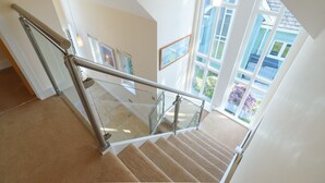 Eine Treppe mit Glasgeländer, die zu einem Balkon mit großen Fenstern und Außenblick führt.