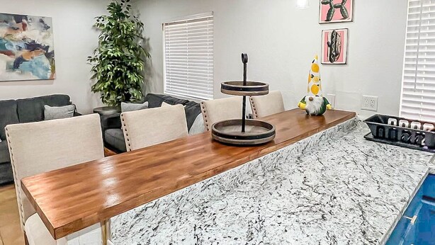 A modern kitchen with a wooden dining table, marble countertops, pendant lights, and a potted plant.