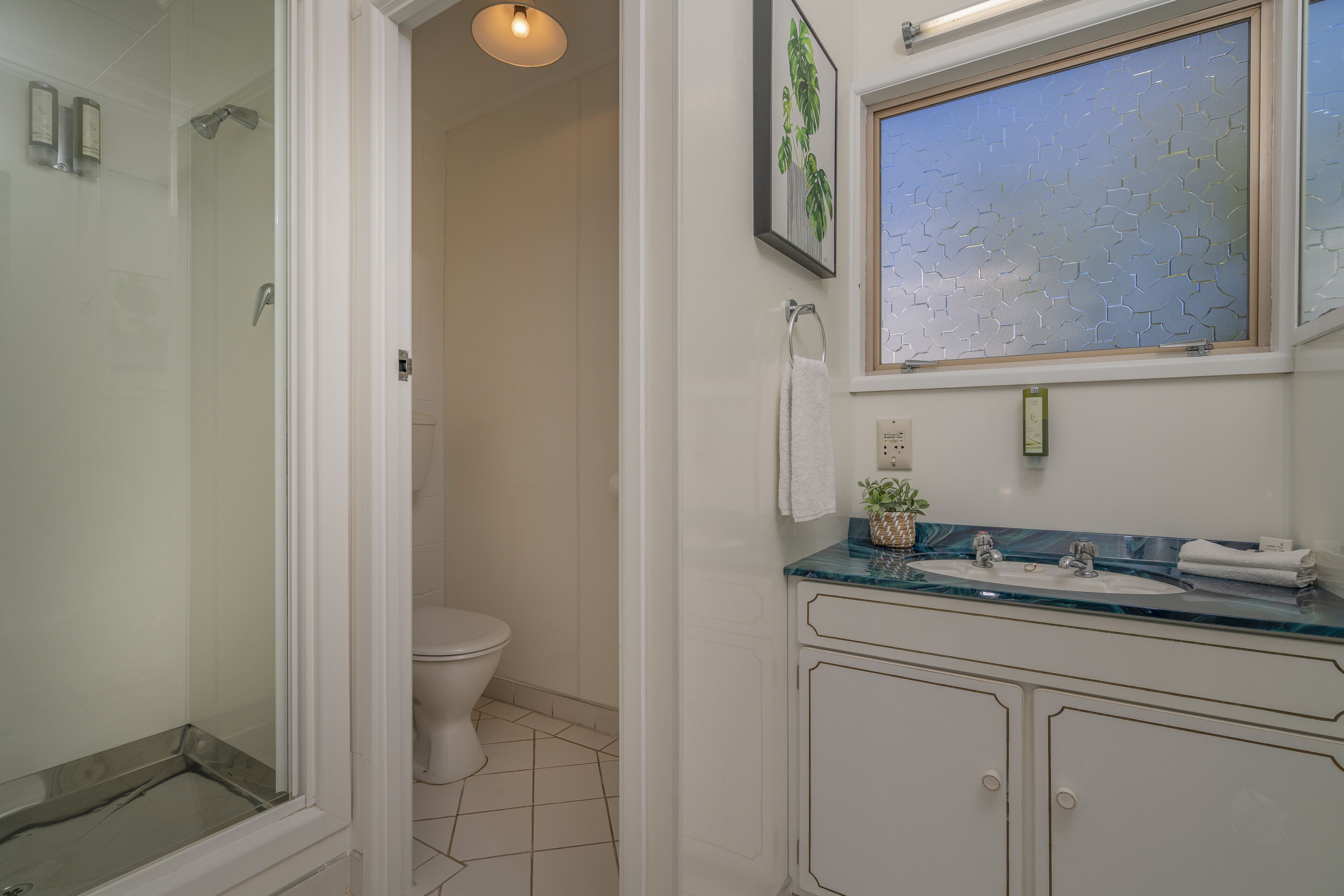 A bathroom with a glass shower enclosure, a toilet, a sink with a marble countertop, and a window with a geometric pattern.