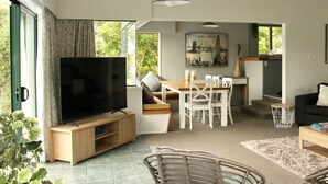 A modern living room with a flat-screen TV, wooden furniture, a dining area, and a large window offering a view of greenery. - Oceans 88 Whitianga Coastal Accommodation Suites (Whitianga)