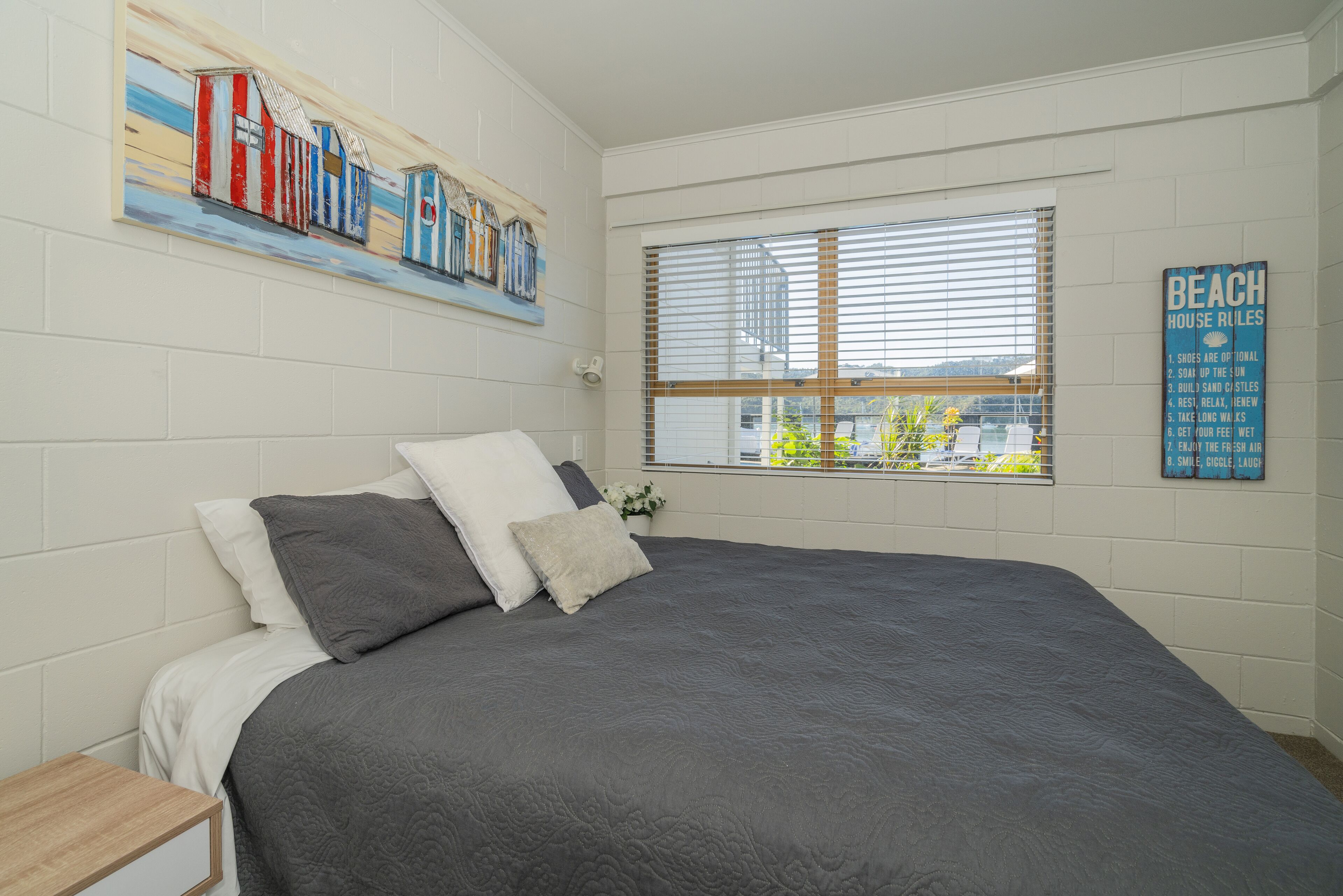 A bedroom with a large bed, a nightstand, a window with blinds, a painting of beach houses, and a sign listing beach house rules.