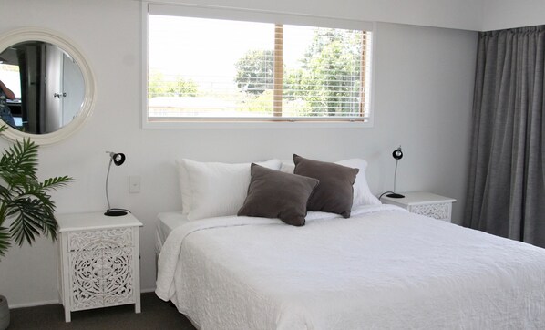 Un dormitorio con cama, dos mesitas de noche, un espejo, una ventana con persianas y una planta.