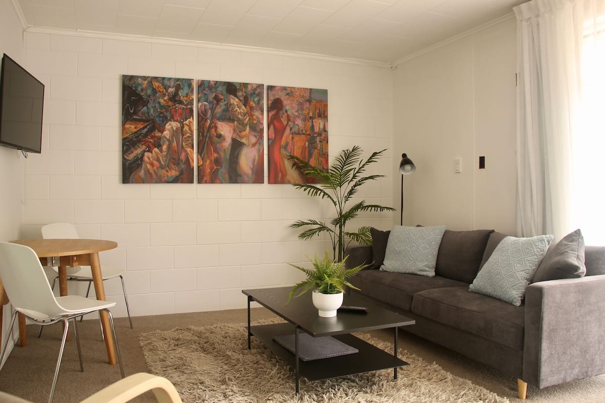 Un salón con sofá, mesa de centro y sillas. Hay dos cuadros en la pared y una planta en maceta.