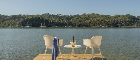 A wooden dock with two white chairs and a small round table with a bottle and flower.