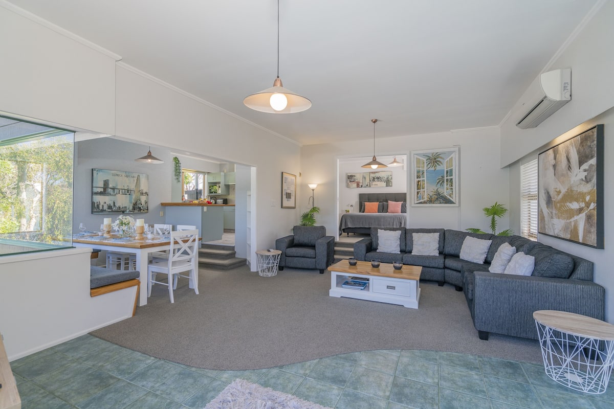 Amplia sala de estar con una mesa de comedor, sofás y una mesa de centro. Se observa un área de cocina al fondo, y la habitación tiene ventanas grandes.