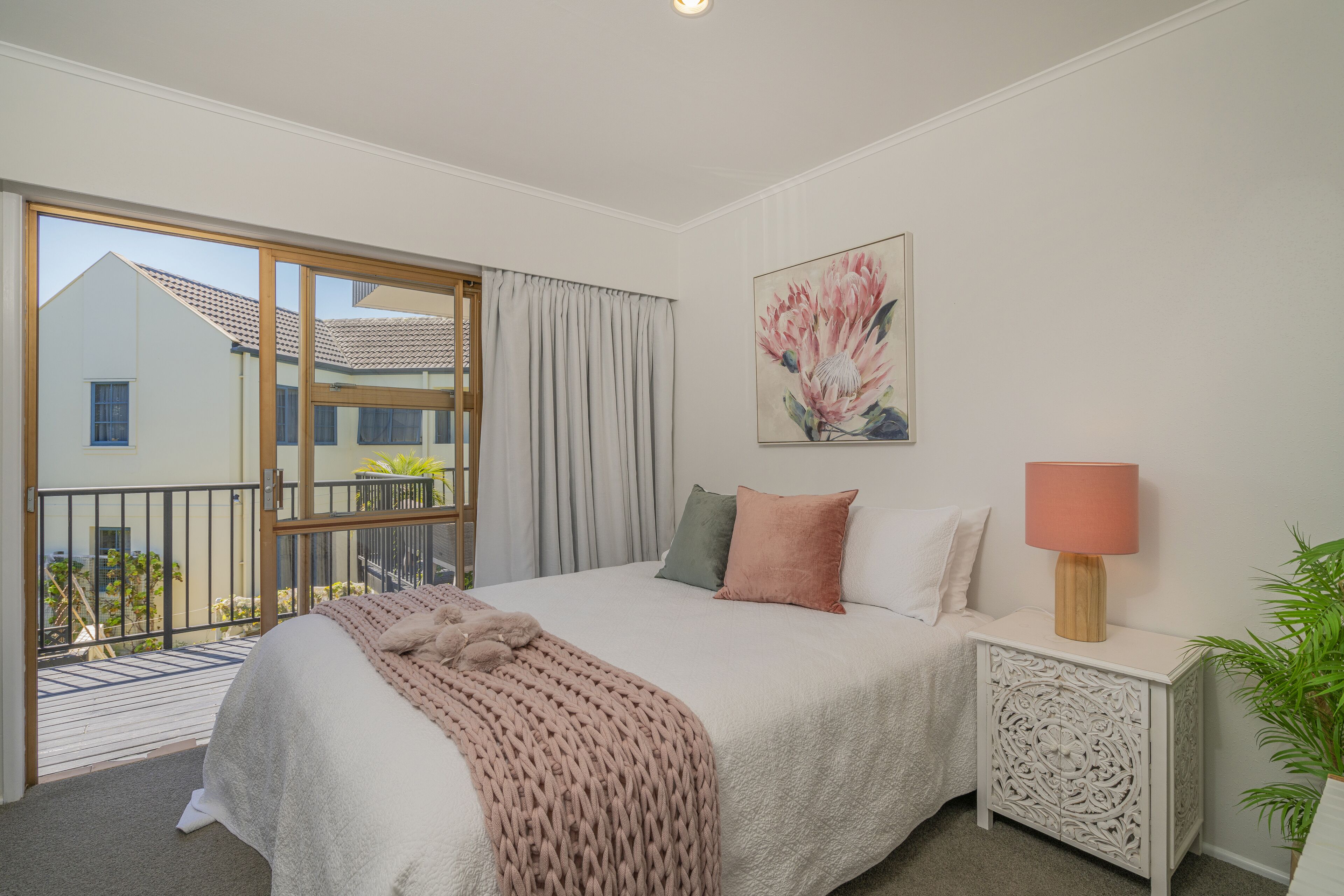 A bedroom with a large bed, a nightstand, a lamp, a painting, and a sliding glass door to a balcony.