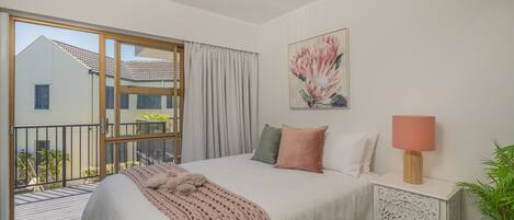 A bedroom with a large bed, a nightstand, a lamp, a painting, and a sliding glass door to a balcony.