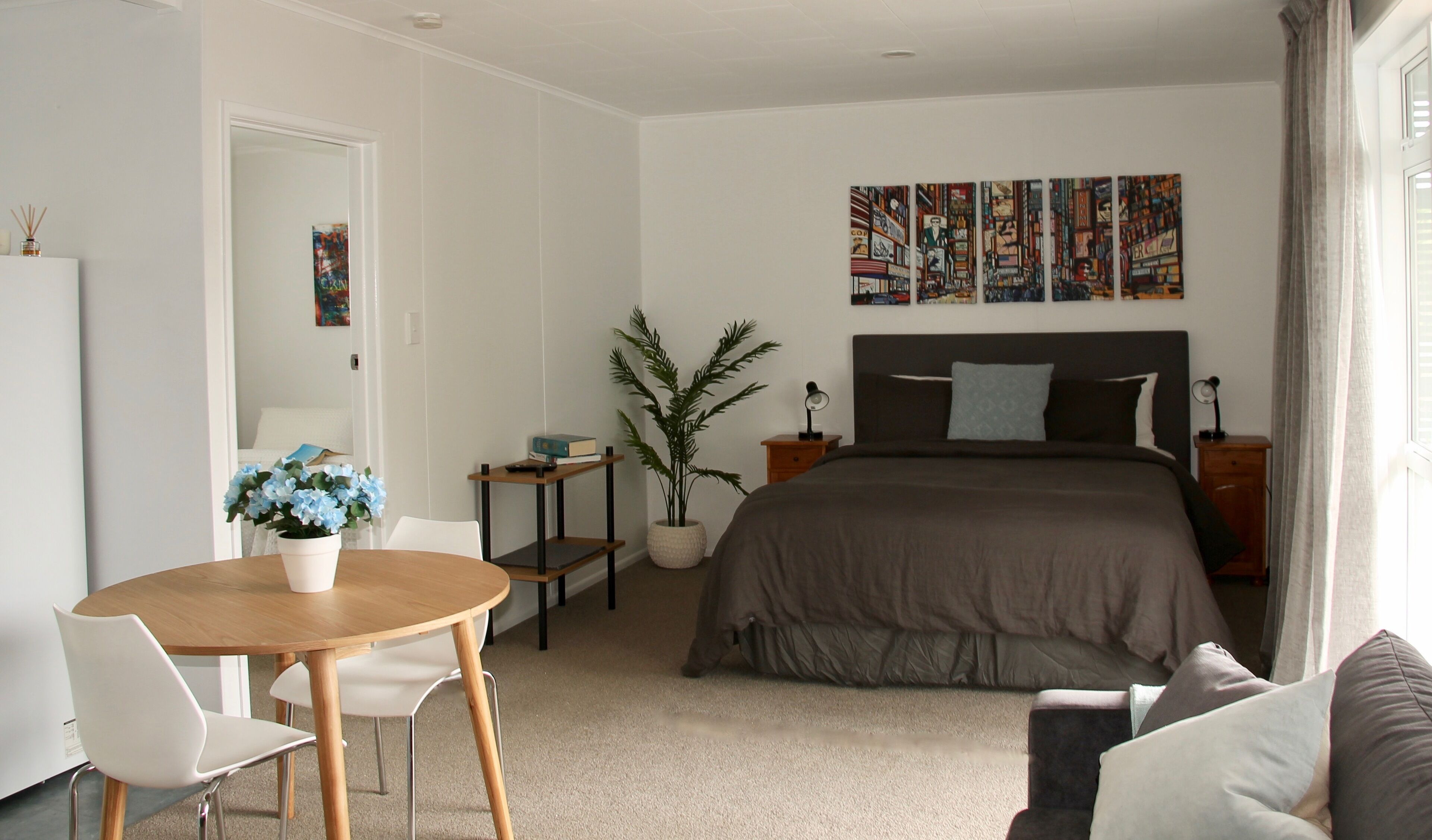 A modern bedroom with a large bed, a bedside table, a nightstand, a desk with a chair, a potted plant, and a wall with framed artwork.