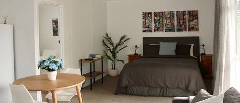 A modern bedroom with a large bed, a bedside table, a nightstand, a desk with a chair, a potted plant, and a wall with framed artwork.