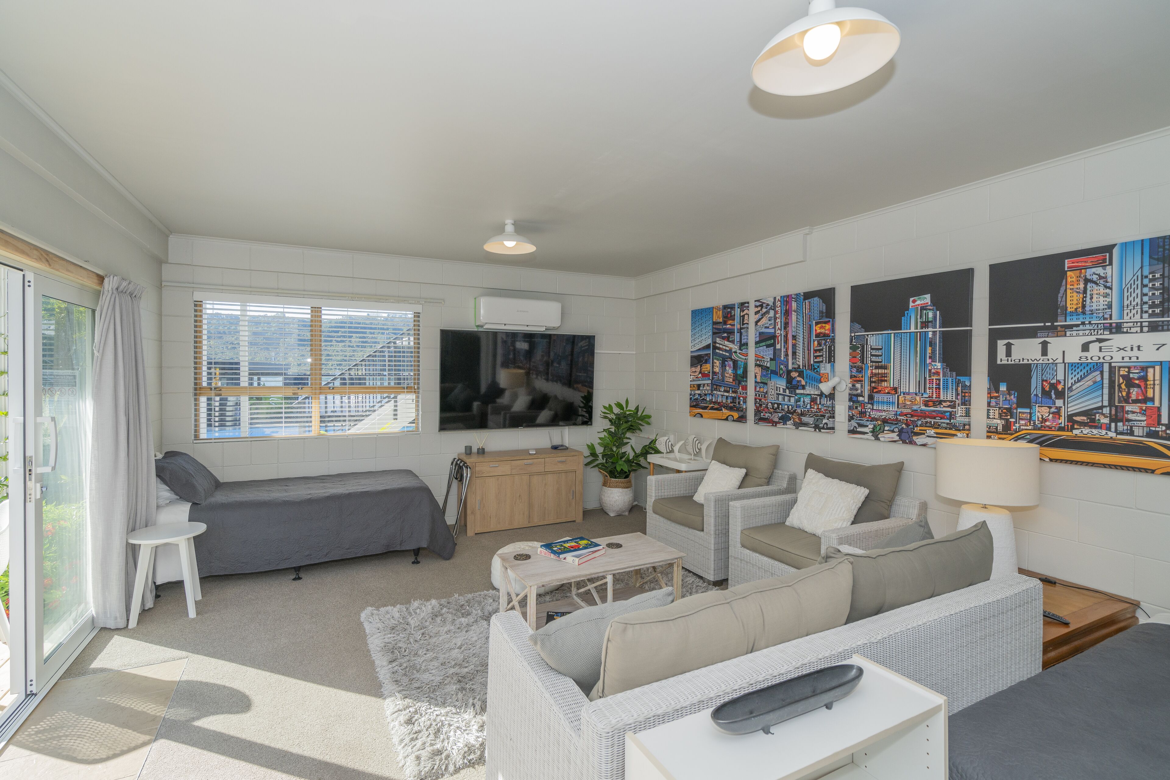 Een moderne woonkamer met een grijze bank, een witte fauteuil, een salontafel en een aan de muur bevestigde televisie. Op de muur hangen stadsgezichtschilderijen en er is een raam met zonwering.