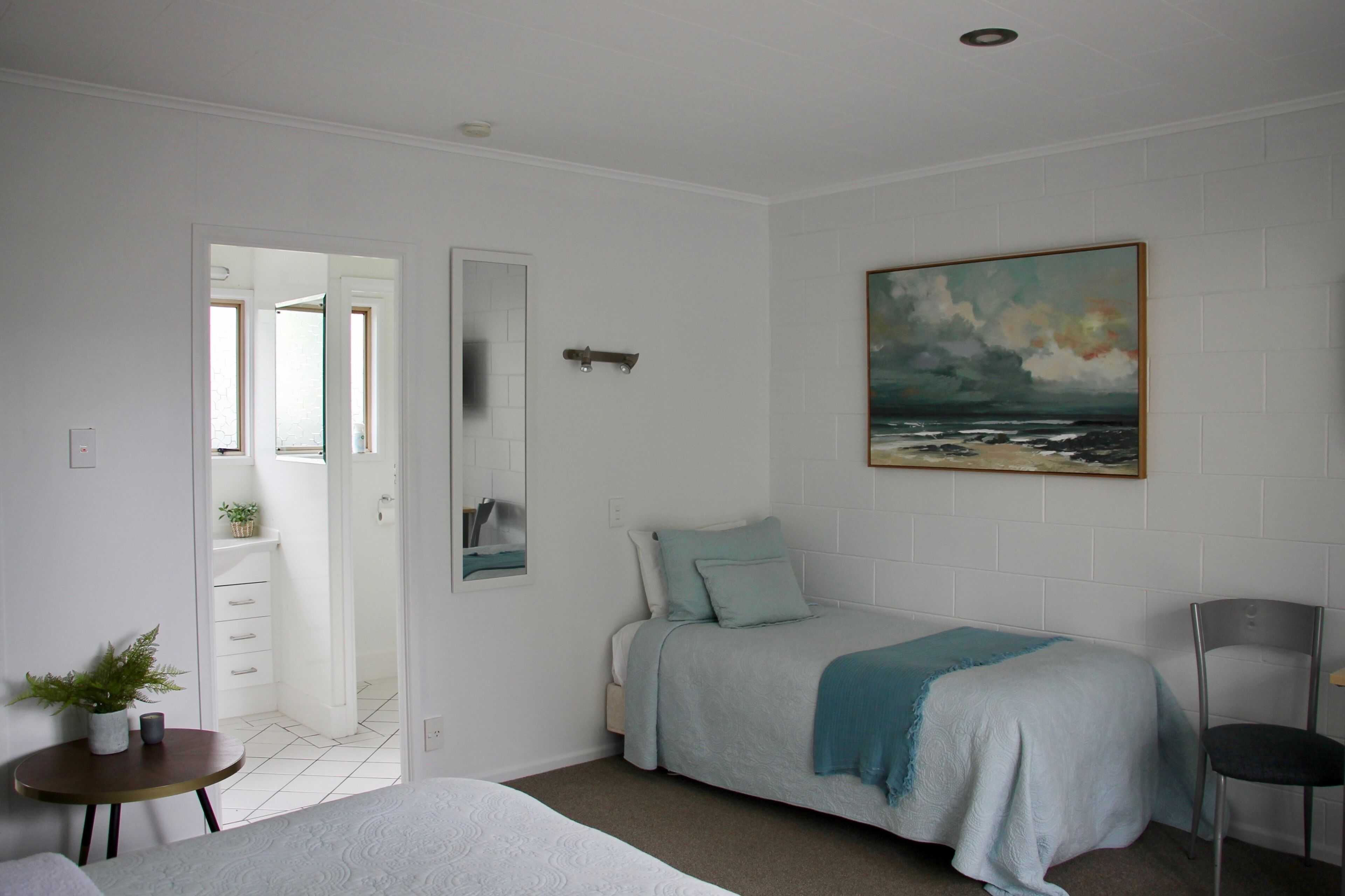 Un dormitorio con cama, una silla, una mesita con una planta y un cuadro de un paisaje marino en la pared.