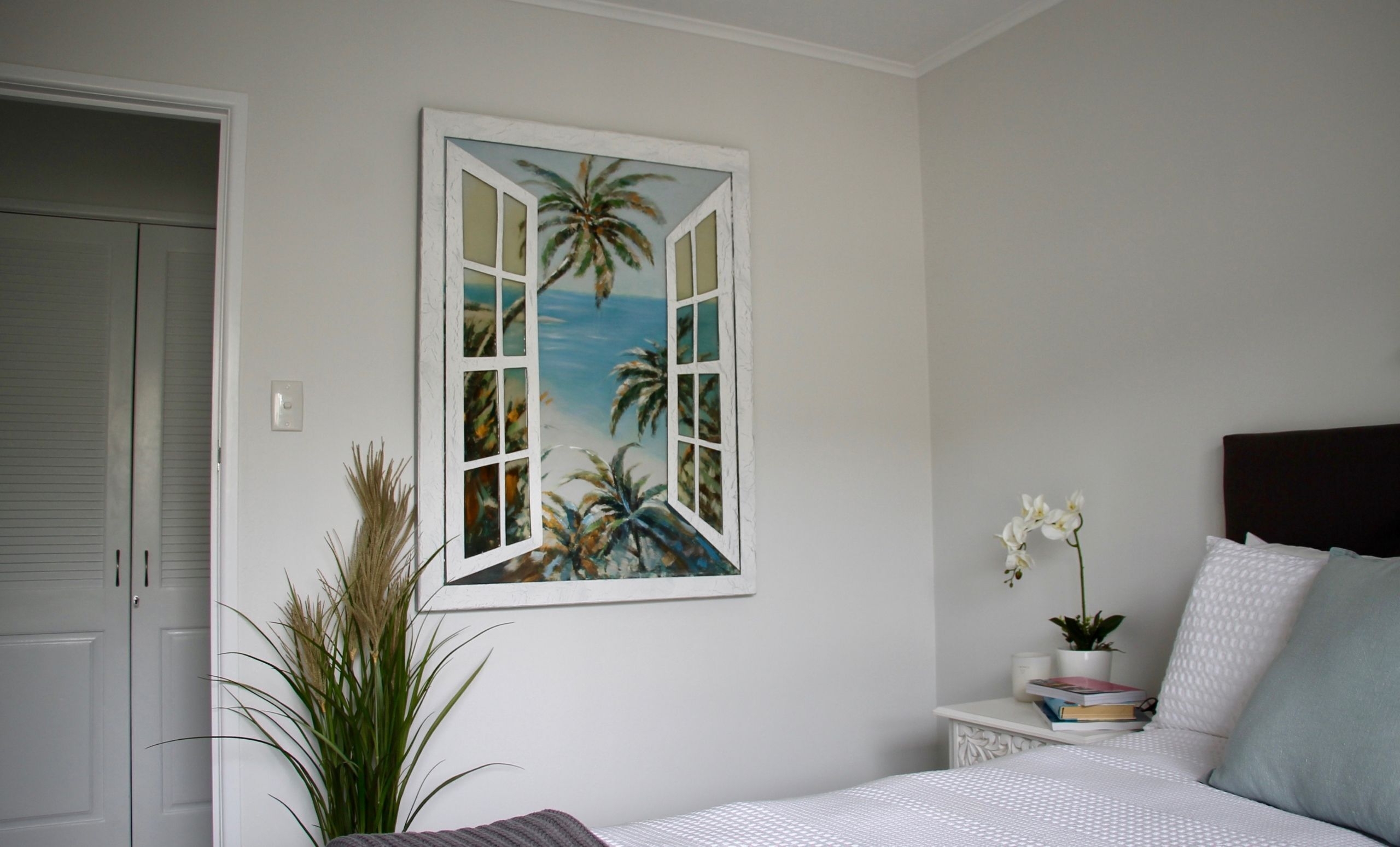 Un dormitorio con una cama, una mesita de noche, una planta en maceta y un cuadro en la pared que representa una ventana con palmeras.