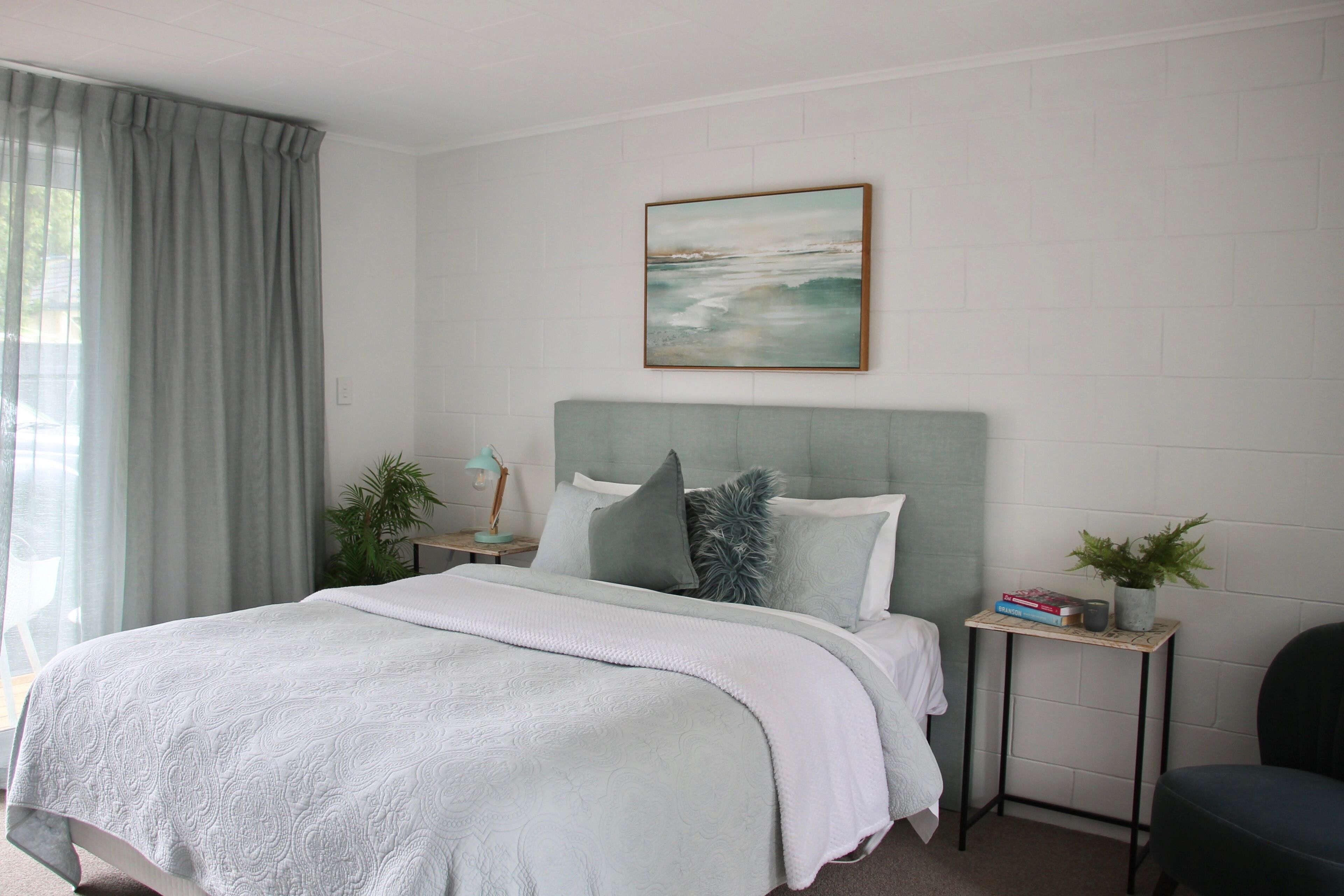 A bedroom with a bed, a bedside table with a lamp, a small plant, and a picture on the wall.