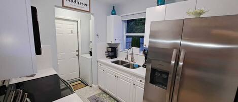 A compact kitchen with white cabinets, a stainless steel refrigerator, a coffee maker, and a window with a view of greenery.