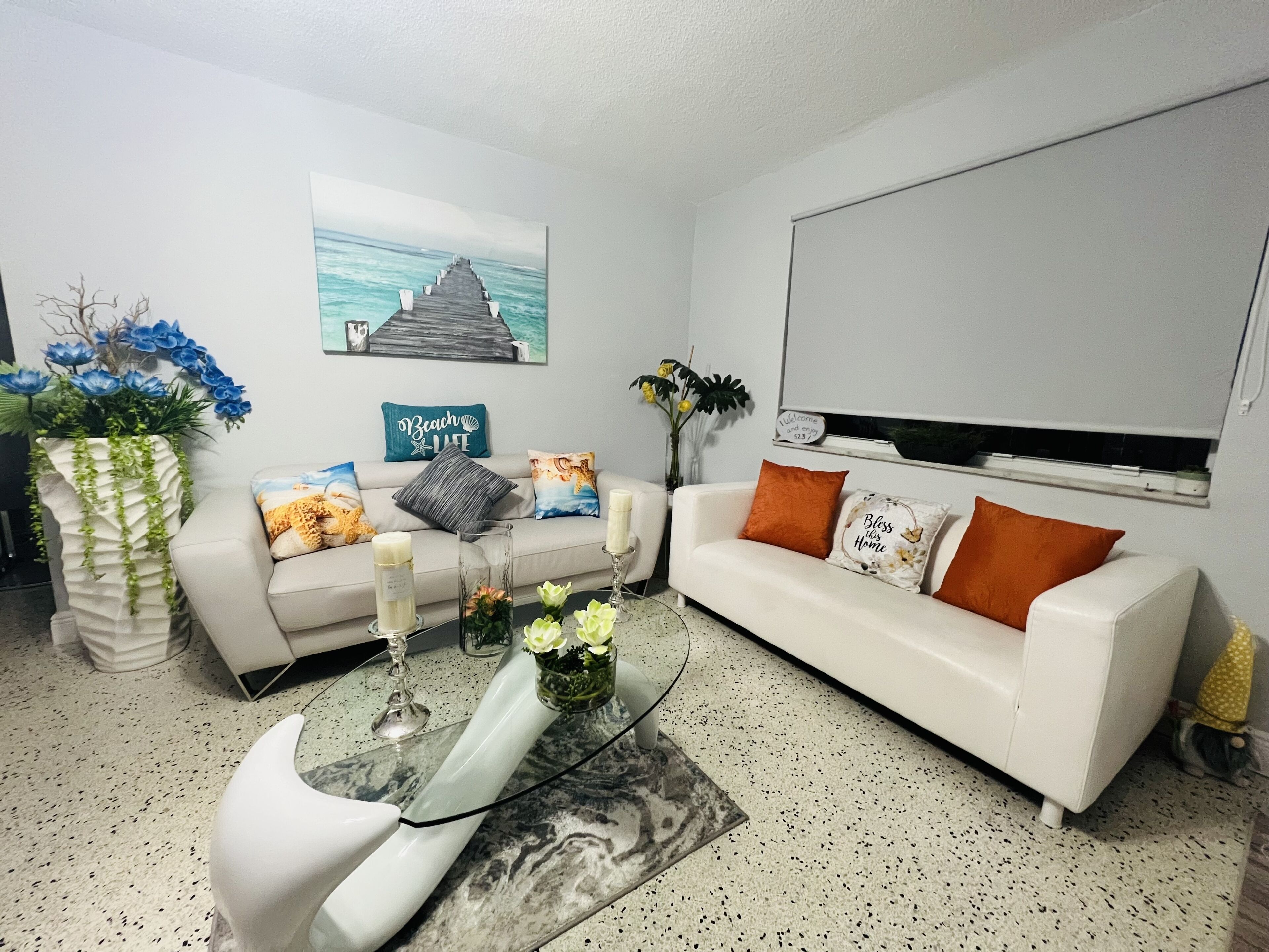 A living room with a white sofa, a coffee table, and decorative pillows. There is a painting on the wall and a small plant on the floor.