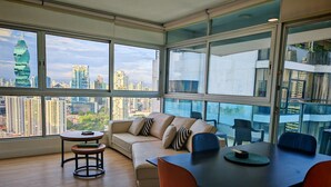 A modern living room with a large window, a beige sofa, a dining table, and orange chairs. - Beutiful High Floor Apartment in the Best Location (Panamá)