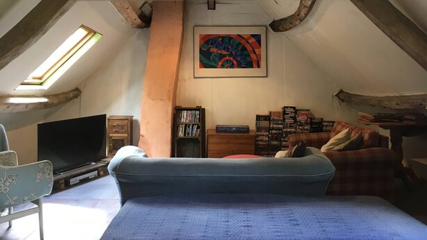 A cozy attic room with a sloped ceiling, a large window, a television, a bookshelf, and a colorful painting. - Charming Rustic 2 bedroom annex cottage (Mouriez)