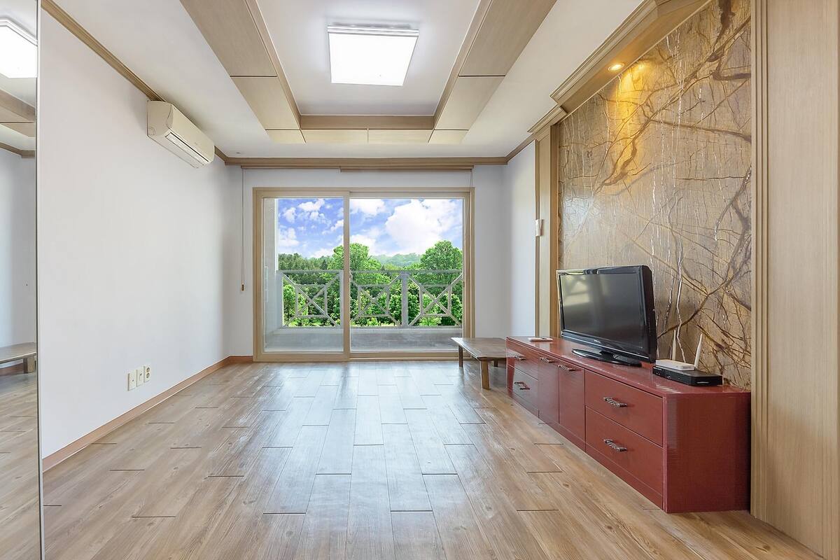 Una habitación amplia con un gran ventanal corredero que da a un balcón, un televisor de pantalla plana sobre un mueble de madera e iluminación empotrada en el techo.