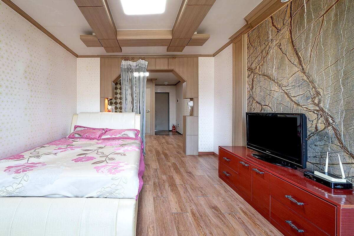 Un dormitorio con una cama, un televisor sobre un mueble de madera, y un pasillo que da a otra habitación.