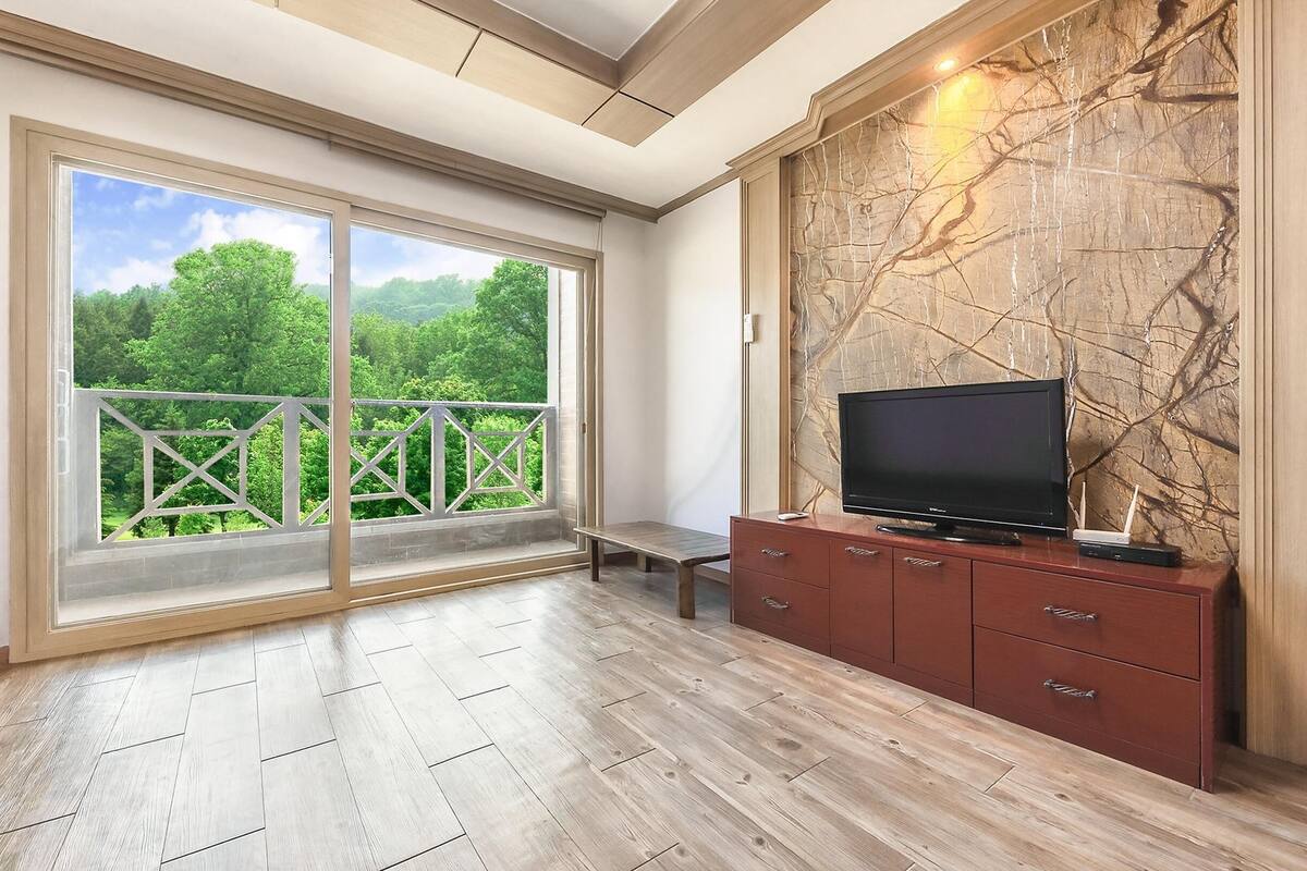 Habitación amplia con un gran ventanal corredera que da a un balcón, un televisor de pantalla plana sobre un mueble de madera y una pared con diseño de mármol.