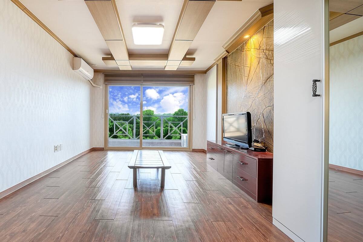 Amplia habitación con piso de madera, un banco, un televisor sobre un mueble de madera y un amplio ventanal corredero que da a un balcón con vistas a zonas verdes.