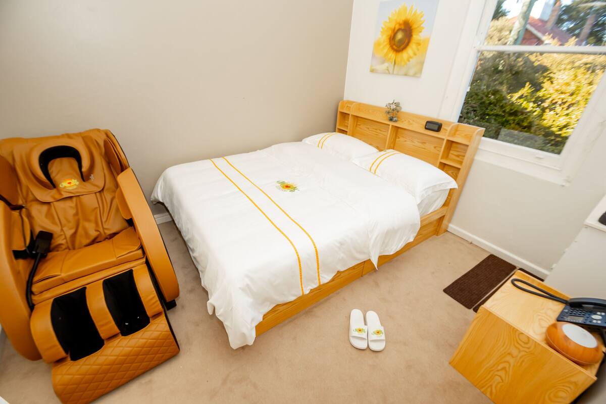 A bedroom with a bed, a massage chair, slippers, a bedside table with a phone, and a window with a view of greenery.