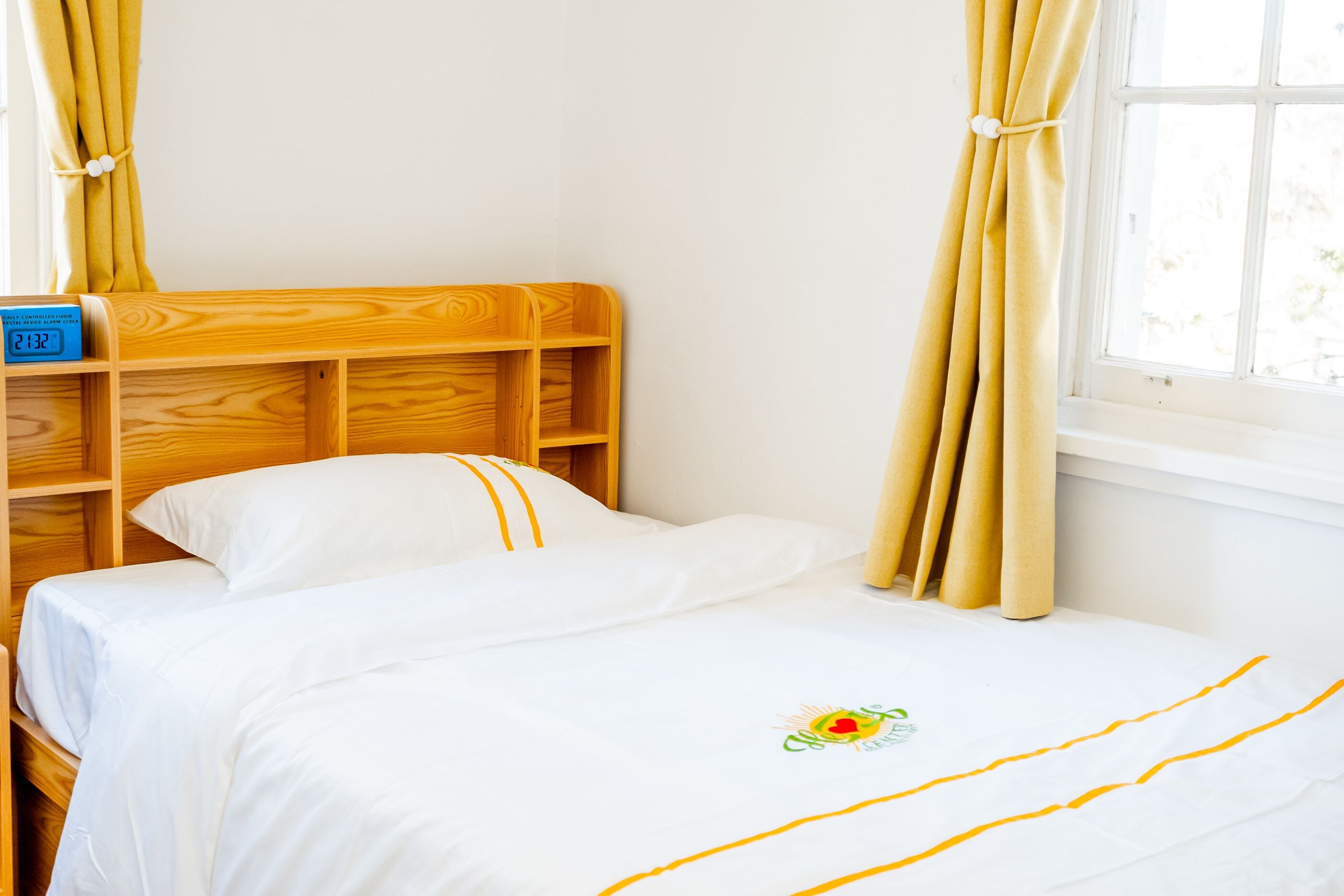 Una cama bien tendida con una colcha blanca y un pequeño logotipo, un cabecero de madera y una ventana con cortinas.