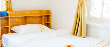 Una cama bien tendida con una colcha blanca y un pequeño logotipo, un cabecero de madera y una ventana con cortinas.