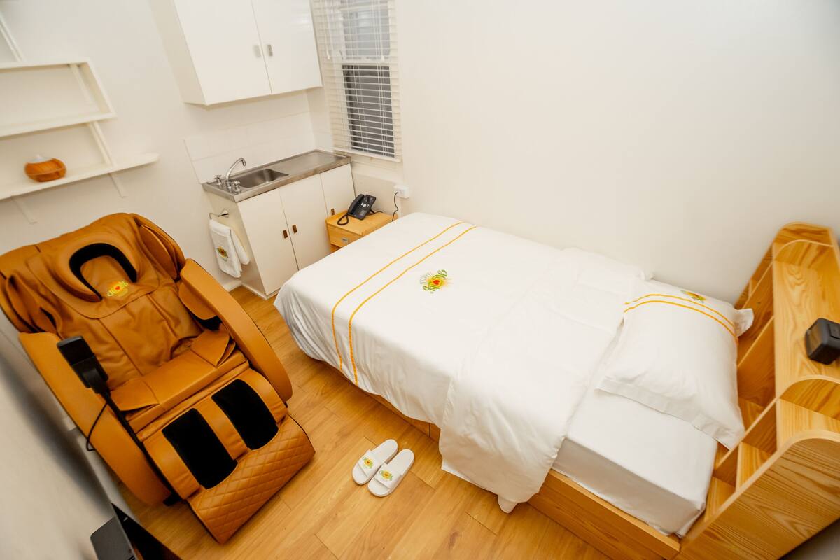 A hotel room with a massage chair, a bed with a white bedspread, a wooden bedside table, a small wooden cabinet, and a window with blinds.