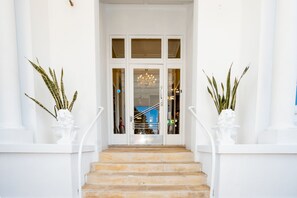 A grand entrance with a staircase leading to a glass door, flanked by white columns and potted plants.