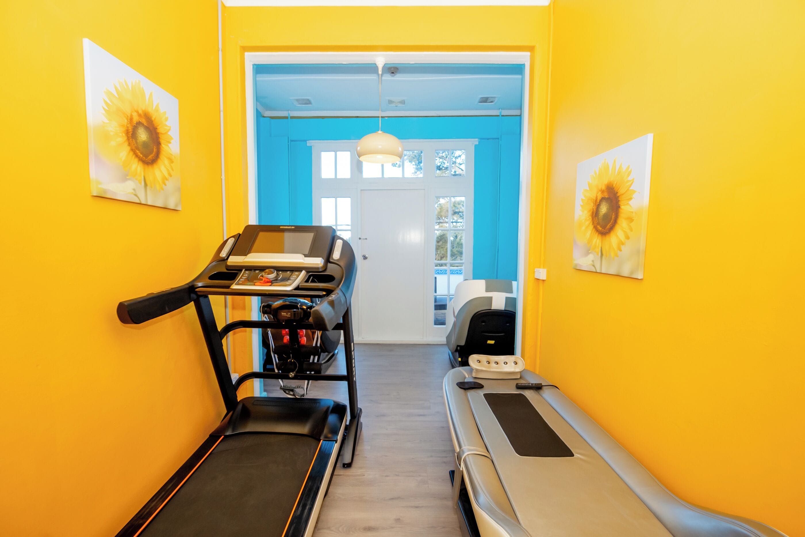 Ein Flur mit Laufband, Ergometer und Massageliege, der zu einer Tür mit blauer Umrandung führt.