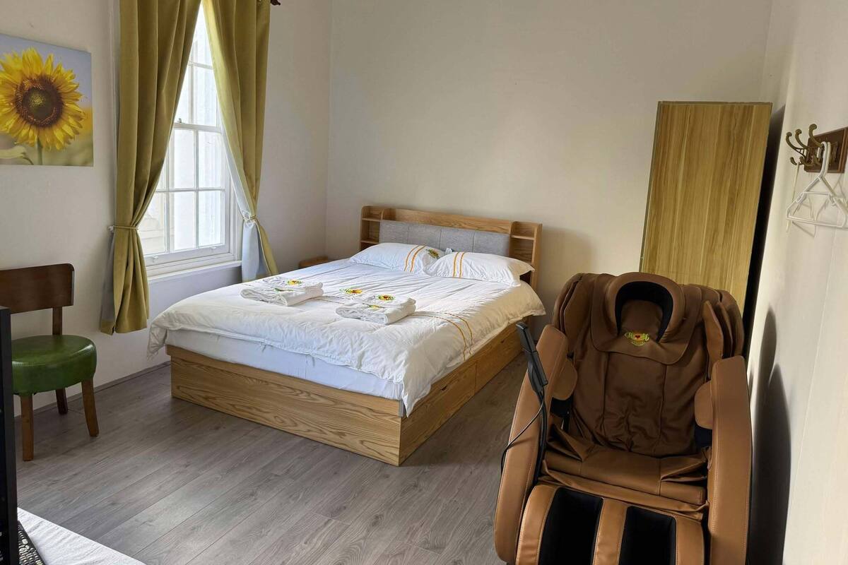 A bedroom with a wooden bed, a green chair, a massage chair, and a large window with curtains.