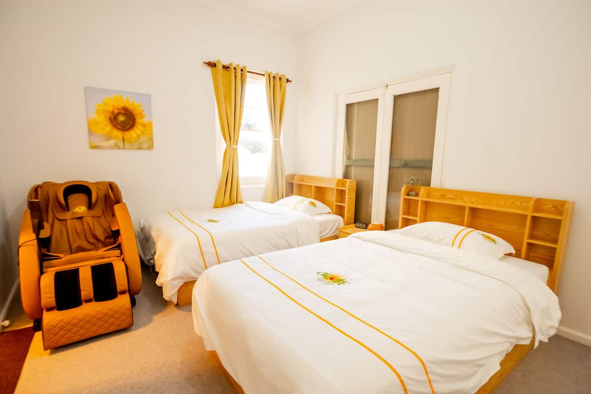 A hotel room with two beds, a massage chair, a window with curtains, and a sunflower painting on the wall.